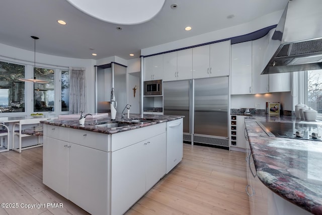 kitchen featuring dark stone counters, light wood-style flooring, built in appliances, a kitchen island with sink, and extractor fan