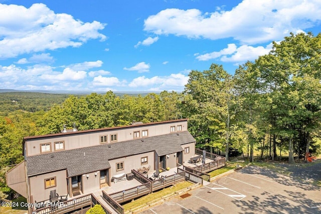 view of front of home with roof with shingles, a wooden deck, uncovered parking, a wooded view, and stucco siding