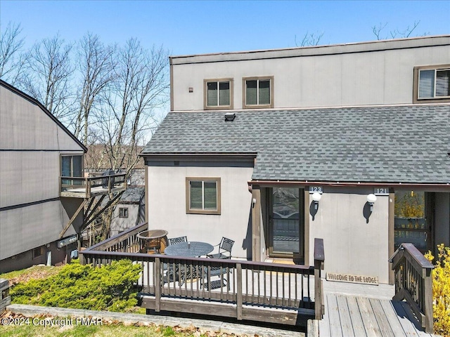 rear view of house with stucco siding, outdoor dining space, a deck, and roof with shingles