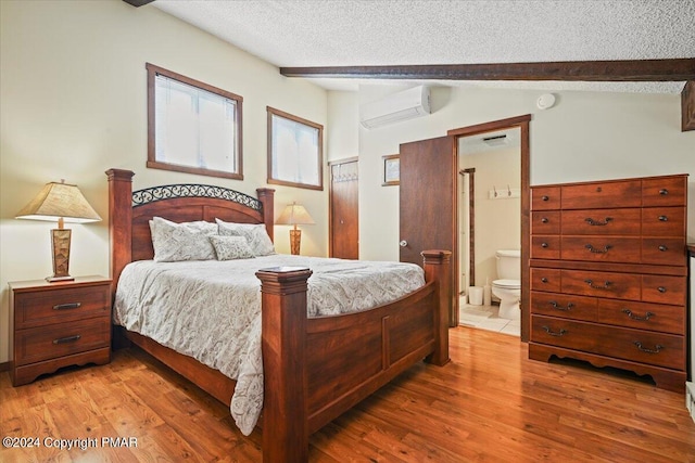 bedroom featuring vaulted ceiling with beams, ensuite bathroom, an AC wall unit, a textured ceiling, and light wood-type flooring