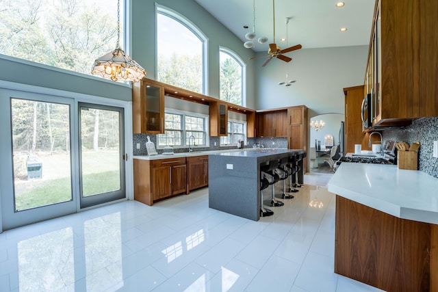 kitchen with brown cabinetry, glass insert cabinets, a sink, ceiling fan with notable chandelier, and backsplash