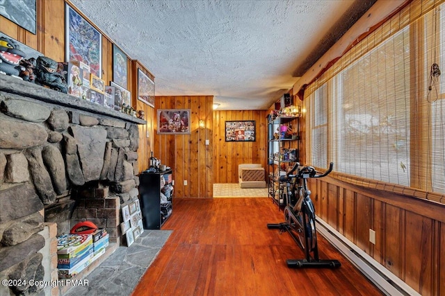 exercise room with wood finished floors, a baseboard heating unit, a stone fireplace, a textured ceiling, and wooden walls