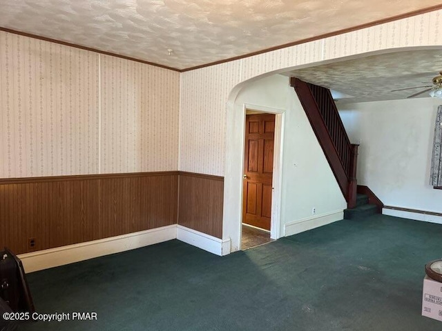 carpeted spare room featuring a ceiling fan, arched walkways, wainscoting, wallpapered walls, and stairs