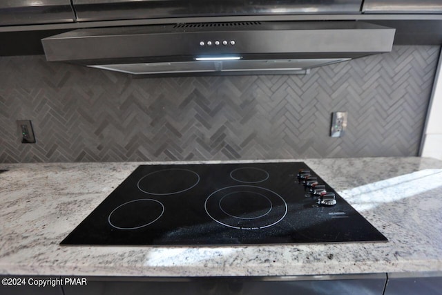 details featuring tasteful backsplash, light stone counters, black electric cooktop, and under cabinet range hood