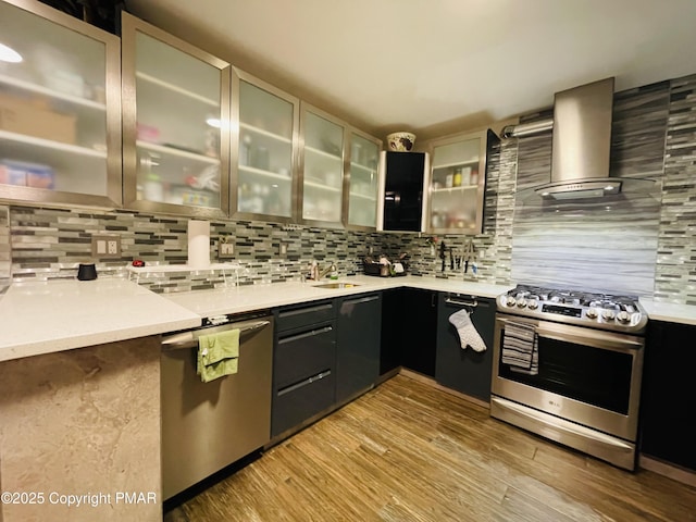 kitchen with light countertops, appliances with stainless steel finishes, wall chimney range hood, tasteful backsplash, and glass insert cabinets