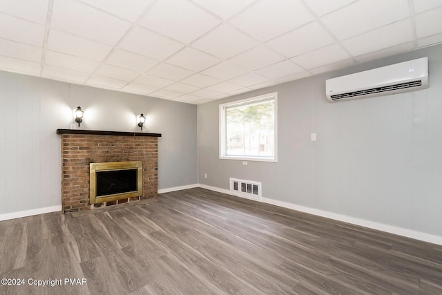 unfurnished living room with visible vents, a drop ceiling, a wall unit AC, wood finished floors, and a brick fireplace