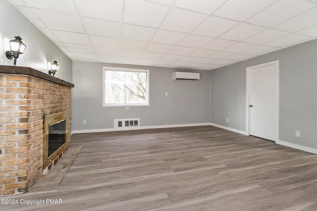 unfurnished living room featuring a drop ceiling, wood finished floors, visible vents, an AC wall unit, and a brick fireplace