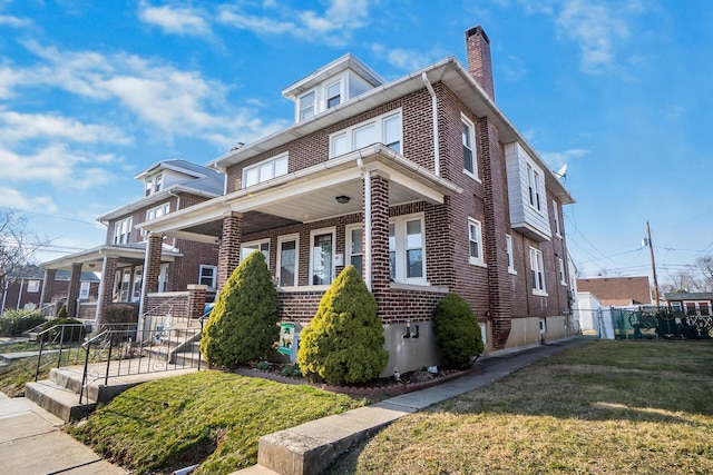 american foursquare style home featuring a porch, fence, a front yard, brick siding, and a chimney