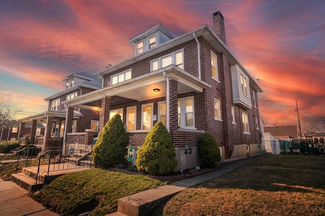 property exterior at dusk with a lawn, fence, covered porch, brick siding, and a chimney