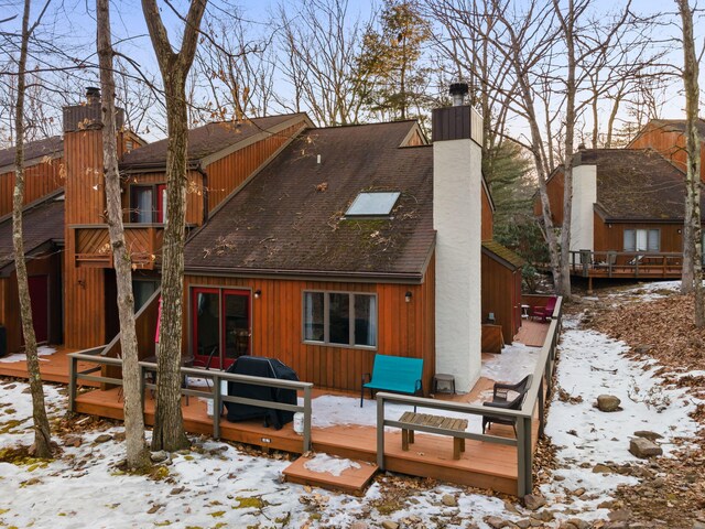 snow covered rear of property with a wooden deck, roof with shingles, and a chimney