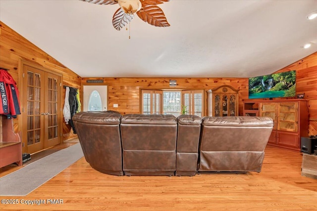 living room with a ceiling fan, light wood-style flooring, vaulted ceiling, french doors, and wood walls