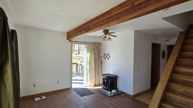 unfurnished room with stairway, a ceiling fan, baseboards, beam ceiling, and a wood stove