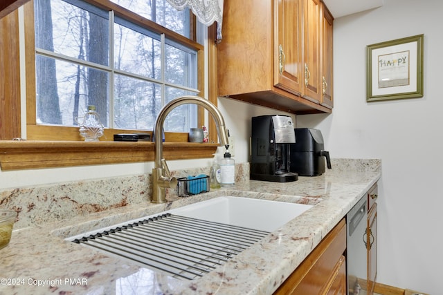 kitchen with brown cabinetry, a sink, light stone counters, and stainless steel dishwasher