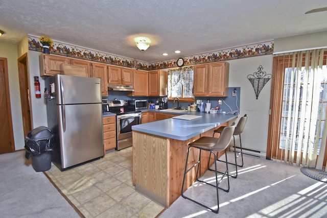 kitchen with a sink, a baseboard heating unit, under cabinet range hood, appliances with stainless steel finishes, and a peninsula