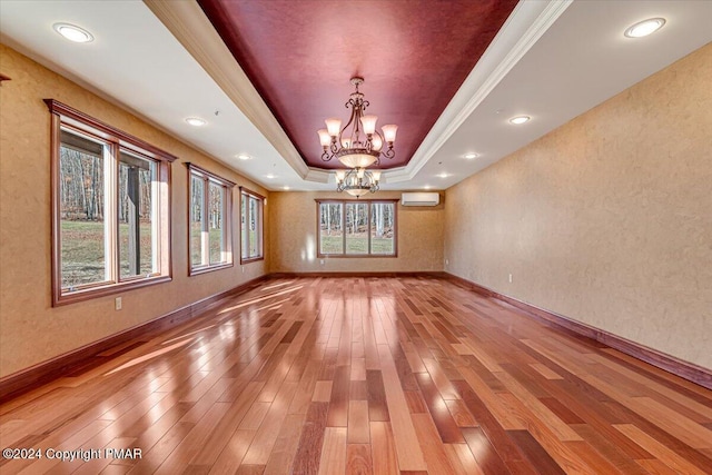 empty room with light wood finished floors, baseboards, a raised ceiling, a wall mounted air conditioner, and a notable chandelier