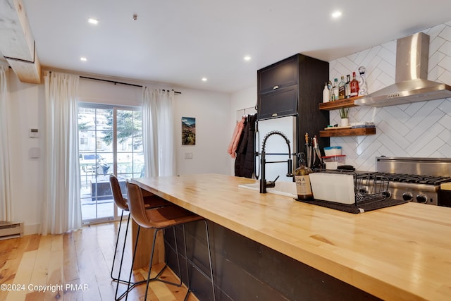 kitchen with wall chimney exhaust hood, light wood finished floors, wooden counters, backsplash, and a kitchen breakfast bar