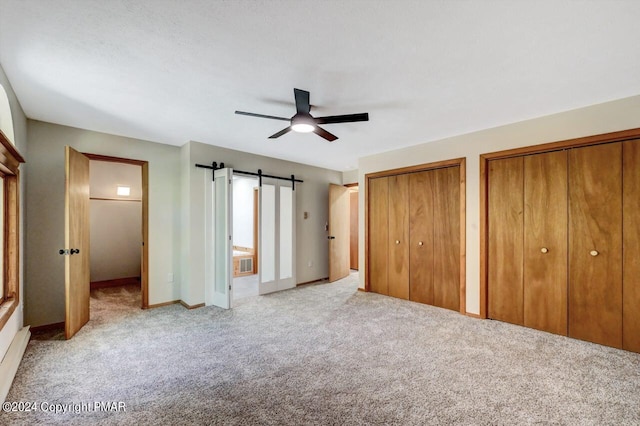 unfurnished bedroom featuring a barn door, a ceiling fan, light colored carpet, baseboard heating, and multiple closets