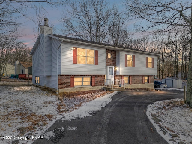 raised ranch featuring a chimney, aphalt driveway, an outbuilding, and brick siding