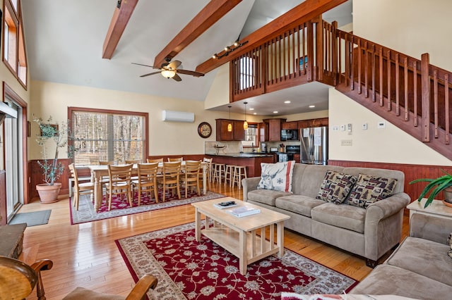 living area featuring light wood finished floors, a wainscoted wall, stairs, beam ceiling, and a ceiling fan