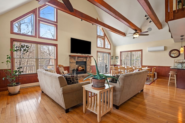 living room featuring a wall unit AC, a ceiling fan, a stone fireplace, light wood-style floors, and wainscoting