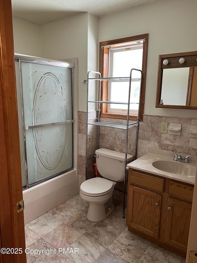 bathroom featuring a textured ceiling, toilet, vanity, tile walls, and combined bath / shower with glass door