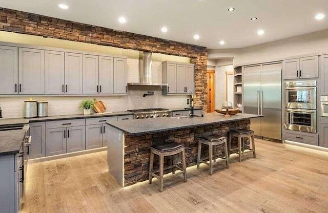 kitchen featuring wall chimney range hood, appliances with stainless steel finishes, a kitchen breakfast bar, recessed lighting, and light wood-style flooring