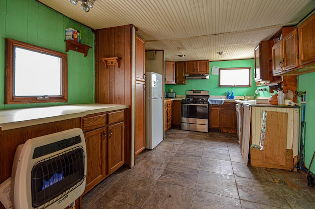 kitchen with heating unit, under cabinet range hood, freestanding refrigerator, brown cabinets, and stainless steel range with gas stovetop