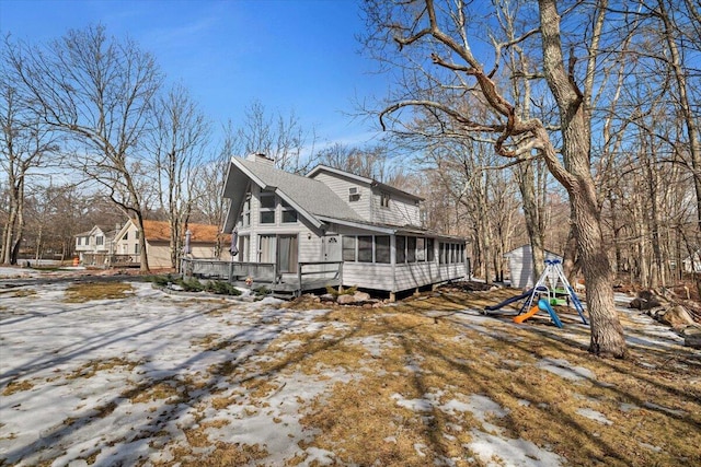 view of snowy exterior with a shingled roof, a sunroom, a playground, and a chimney