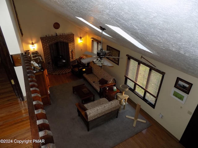 living room featuring dark wood-type flooring, lofted ceiling with skylight, a brick fireplace, a textured ceiling, and ceiling fan