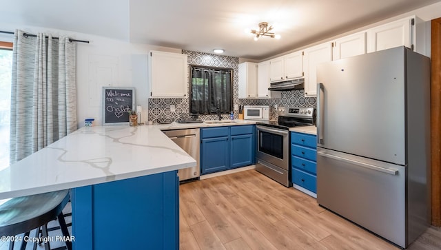 kitchen with a peninsula, blue cabinetry, under cabinet range hood, appliances with stainless steel finishes, and white cabinetry