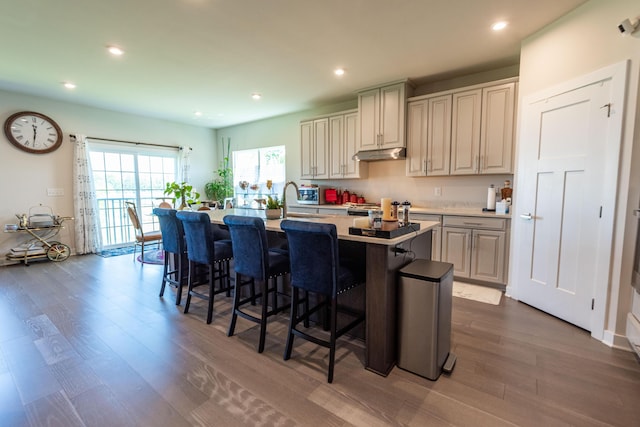 kitchen with dark wood-style floors, under cabinet range hood, a center island with sink, a kitchen bar, and recessed lighting