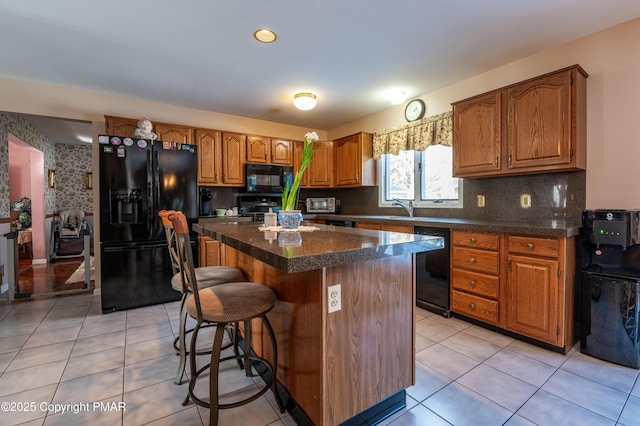 kitchen with dark countertops, brown cabinetry, a sink, a kitchen island, and black appliances