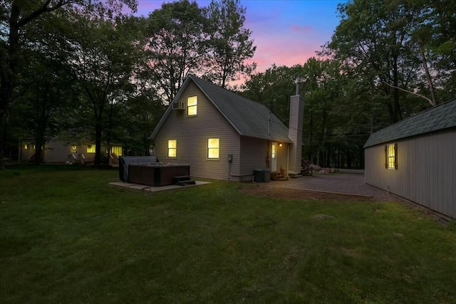 back of property at dusk featuring a yard, a chimney, a patio area, and a hot tub