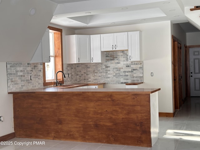 kitchen with a raised ceiling, backsplash, white cabinets, a sink, and a peninsula