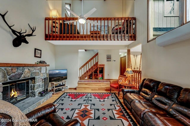 living area featuring stairs, a stone fireplace, a towering ceiling, wood finished floors, and a ceiling fan