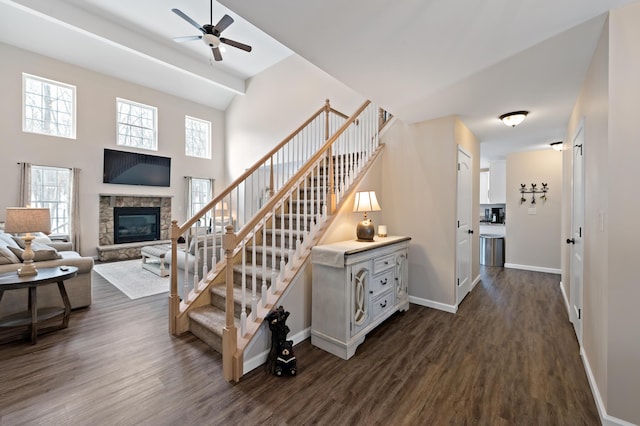 staircase featuring a high ceiling, ceiling fan, a stone fireplace, wood finished floors, and baseboards