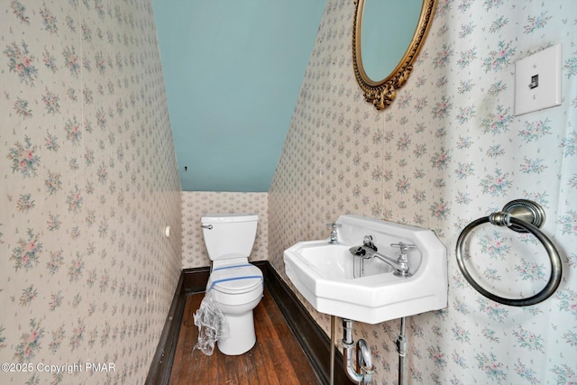 half bathroom with toilet, wallpapered walls, a sink, and wood finished floors