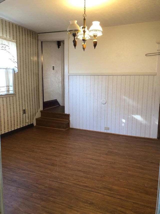 empty room with wood finished floors, a wainscoted wall, wallpapered walls, an inviting chandelier, and stairs