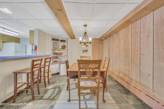 dining area featuring baseboard heating, a drop ceiling, wood walls, and a notable chandelier