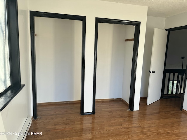 unfurnished bedroom with a textured ceiling, two closets, a baseboard heating unit, and wood finished floors