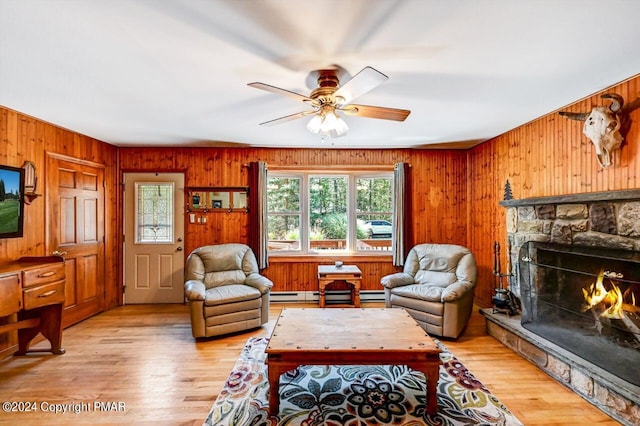 living area featuring a baseboard radiator, light wood-style flooring, ceiling fan, a stone fireplace, and wooden walls