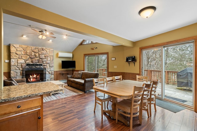 dining area with a wainscoted wall, vaulted ceiling with beams, a stone fireplace, wood finished floors, and a wall mounted air conditioner