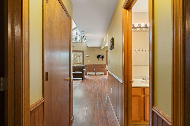 hallway with a baseboard radiator, dark wood-type flooring, wood walls, a sink, and wainscoting