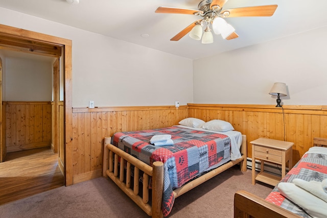 carpeted bedroom featuring wood walls, a baseboard radiator, a ceiling fan, and wainscoting