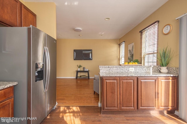 kitchen with stainless steel fridge, brown cabinetry, wood finished floors, a peninsula, and light stone countertops