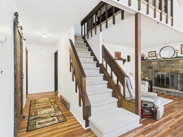 staircase with a barn door, baseboard heating, wood finished floors, and a stone fireplace