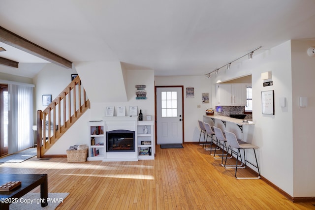 foyer entrance featuring baseboards, a glass covered fireplace, stairway, light wood-style floors, and beam ceiling