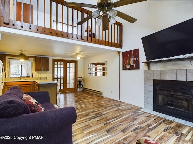 living room with french doors, a towering ceiling, baseboard heating, wood finished floors, and a tile fireplace