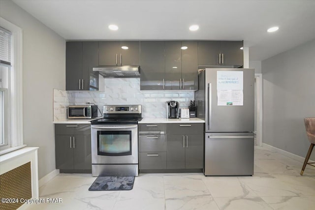 kitchen featuring marble finish floor, tasteful backsplash, gray cabinetry, appliances with stainless steel finishes, and under cabinet range hood