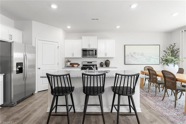 kitchen with appliances with stainless steel finishes, wood finished floors, a kitchen island with sink, white cabinets, and light stone countertops